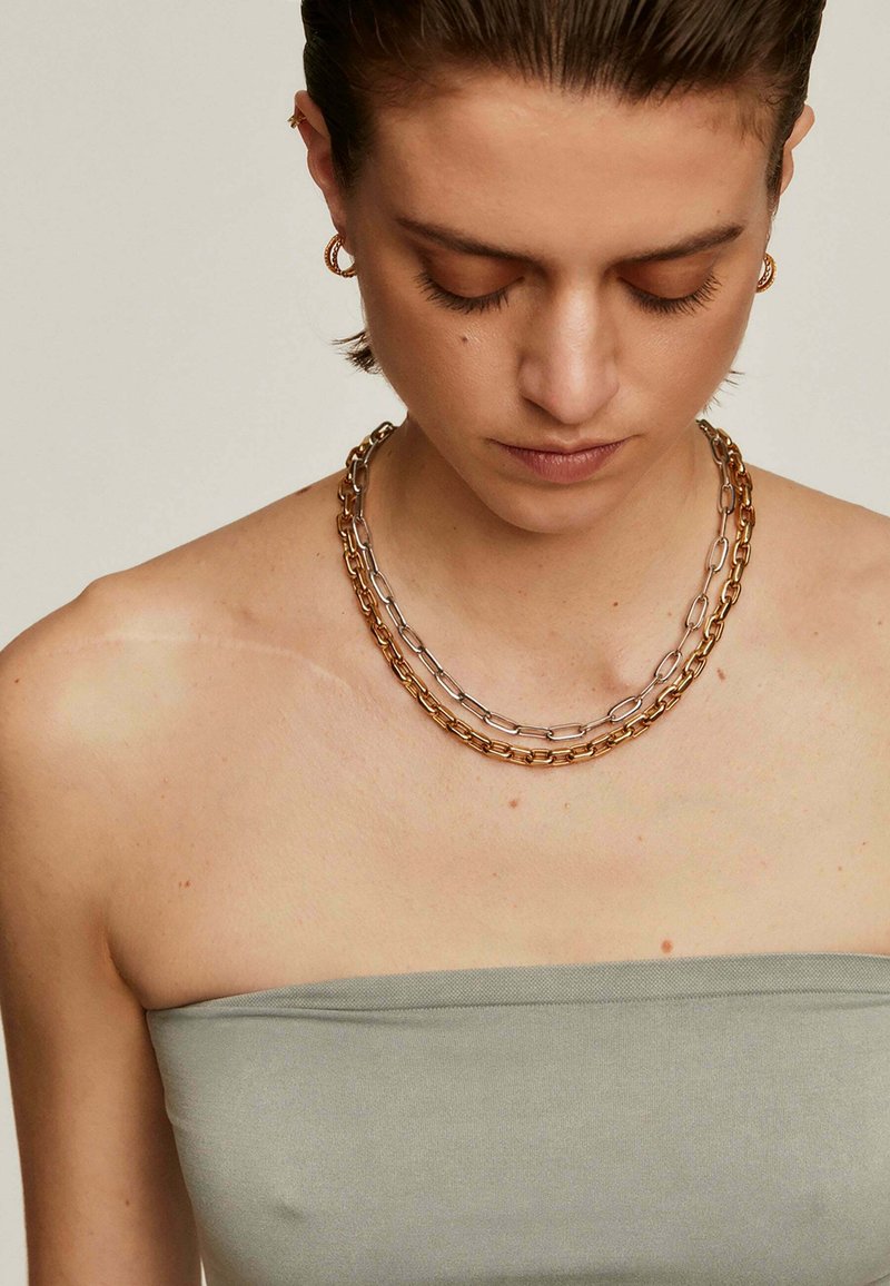 Collar de cadena con eslabones de plata y oro en capas, con una textura suave. Lleva puesto en un hombro descubierto.