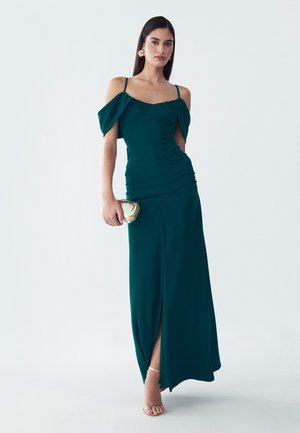 Mujer con vestido de noche verde azulado con los hombros al descubierto y abertura, sosteniendo un bolso de mano dorado, llevando sandalias doradas y pendientes en forma de hoja sobre un fondo liso.