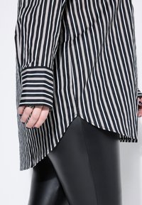 Camisa larga de rayas blancas y negras con dobladillo curvado, que cuenta con mangas largas y una textura suave, combinada con leggings negros.