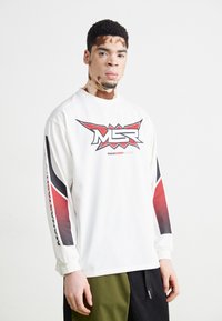 Weißes Langarmhemd mit einem grafischen Logo in Rot und Schwarz. Verfügt über gemusterte Akzente an den Ärmeln und einen lockeren Schnitt. Kombiniert mit grünen Shorts.