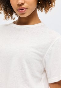 Νεαρό άτομο με σγουρά μαλλιά που φοράει ένα απλό λευκό μπλουζάκι με κοντά μανίκια και χρυσό σκουλαρίκι κρίκο, με τα χείλη ελαφρώς ανοιχτά.