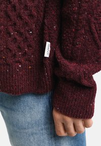 Suéter de punto burdeos con patrón de trenzado texturizado, puños acanalados y una etiqueta blanca que presenta "GANT". Lleva puesto sobre jeans de denim azul.