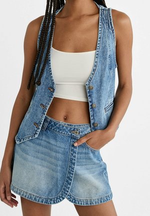 Vrouw met lichtblauw denimvest en wikkelrok van denim over een witte cropped tanktop, hand in de zak van de rok.