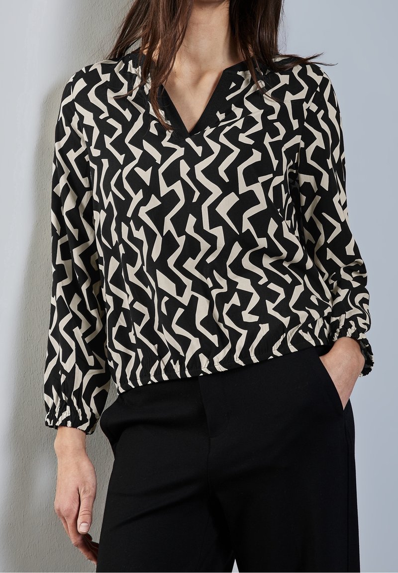 Blouse noire et crème à motifs avec un col en V, des poignets élastiques et une taille ajustée. Le motif présente des formes abstraites en zigzag.