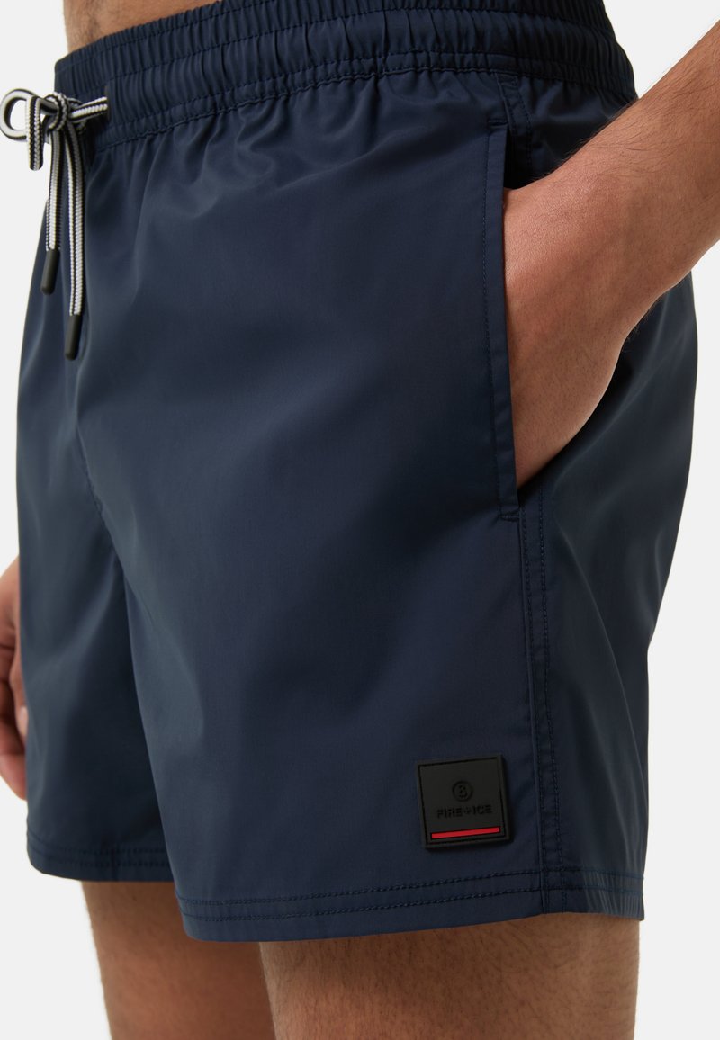 Marineblaue Schwimmshorts mit elastischem Bund, Kordelzug, Seitentaschen und einem Logopatch mit roten und schwarzen Akzenten. Glattes, mattes Material.