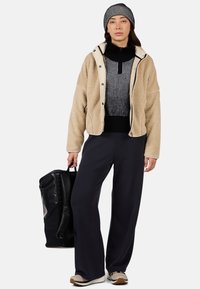 Beige, strukturierte Fleecejacke über einem schwarz gestreiften Pullover; weite schwarze Hosen; gestreifte Mütze. Hält eine schwarze Tasche und trägt beige Sneakers.
