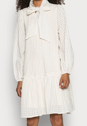 Kobieta w białej sukience do kolan z długimi bufkami, wzorem z otworków w kwadratowy wzór oraz dużą kokardą zawiązaną na szyi.