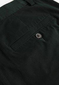 Donkergroene broek van gladde stof, met een achterzak voorzien van een enkele bruine knoop en gestikte details langs de naden.