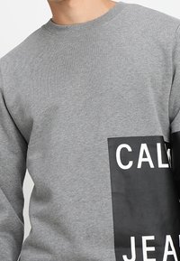 Šedá mikina vyrobená z bavlněné směsi, s velkým černým grafickým panelem a bílým textem "CALM JEANS" na levé straně.