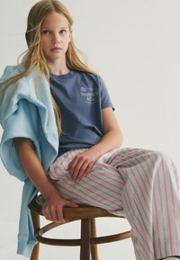 Námořnické tričko s logem "Superdry & Co", světle modrá mikina přes jedno rameno a růžové pruhované pyžamové kalhoty se zelenými pruhy.