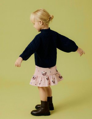 Pull en maille bleu marine avec des manches ballon, associé à une jupe rose à volants comportant des motifs de cônes de glace. Bottines à talons marron foncé.