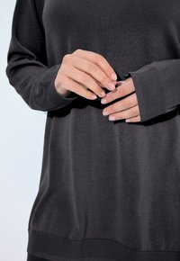 Mains avec des ongles naturels ajustant les manches longues d'un pull sombre gris contre un fond clair.