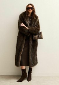 Paltò in pelliccia sintetica marrone con colletto ampio, abbinato a un vestito a coste scuro e stivaletti marroni. Gli accessori includono una borsa testurizzata e occhiali da sole.