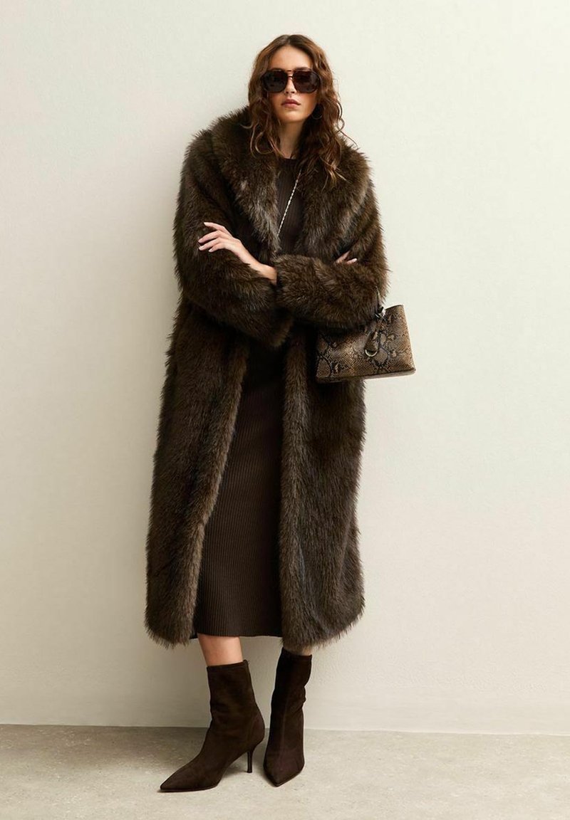 Paltò in pelliccia sintetica marrone con colletto ampio, abbinato a un vestito a coste scuro e stivaletti marroni. Gli accessori includono una borsa testurizzata e occhiali da sole.