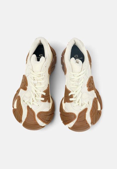 Een paar witte sneakers met dikke bruine rubberen zolen met een getextureerd, segmentontwerp, gezien van bovenaf op een witte achtergrond.