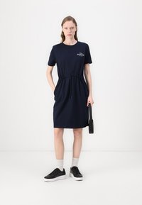 Femme portant une robe bleu marine à manches courtes avec cordon à la taille, tenant un sac à main noir, des chaussettes blanches et des baskets noires.