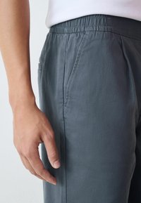 Könnyű, sötétszürke szövetnadrág, rugalmas derékkal, oldalsó zsebekkel és sima felülettel, egy kéz a szövetet érintve.