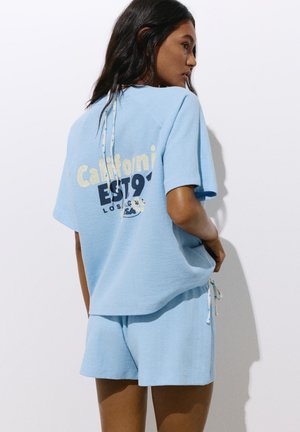 Femme portant un short tricoté bleu clair et une chemise ample avec le texte "California EST 1978" dans le dos, debout contre un mur blanc.