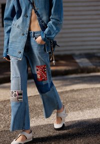 Denim-yhdistelmä, johon kuuluu takki ja leveälahkeiset farkut, joissa on päällysteitä, värikkäitä yksityiskohtia ja erilaisia tekstuureja. Esillä valkoiset kengät.