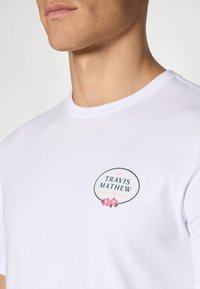 Kurzärmliges weißes T-Shirt aus Baumwolle mit einem runden Logo "TRAVIS MATHEW" und floralen Akzenten auf der linken Brustseite.