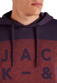 Sudadera con capucha en morado oscuro y rojo, con un patrón texturizado que presenta las letras en negrita "JACK" y "&". Cuerdas de ajuste con puntas metálicas.