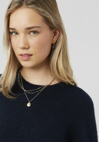 Pull marine foncé avec un col rond. Chaînes en or superposées, l'une avec un pendentif rond, et de petites boucles d'oreilles en forme de compas noir.