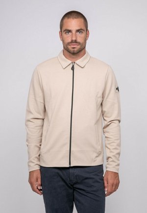 Homme aux cheveux courts portant une veste zippée beige et un pantalon foncé, debout devant un fond uni gris clair, faisant face à l'avant.