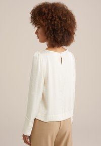 Blusa bianca, texture, con maniche lunghe, scollatura rotonda e dettaglio a chiave sul retro, abbinata a pantaloni beige. Design semplice ed elegante.