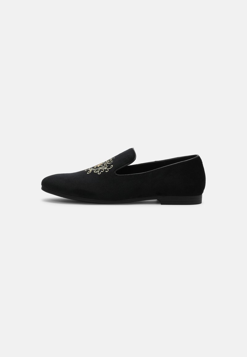 Chaussure noire en velours à enfiler, à petit talon et avec un écusson décoratif brodé en or sur la partie supérieure.