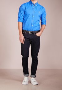 Ljusblå skjorta med knappar, mörka jeans, svart bälte och vita sneakers med uppvikta fållar. Fokus på färg och tygstruktur.