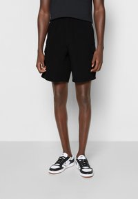 Person som bär svarta cargoshorts och svartspräckliga Vans-sneakers, stående framför en enkel ljusgrå bakgrund.