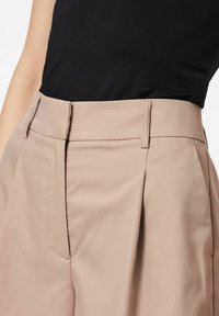 Pantalons beiges taille haute avec des plis, en tissu lisse. Présente un devant plat et des poches latérales, associés à un haut noir.