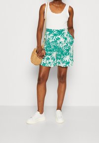 Zelená a bílá sukni se vzorem tropických listů s kapsami, doplněná bílým ribkovaným topem a bílými teniskami. Texturovaná slámová kabelka.