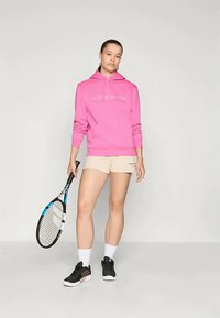 Ružičasta hoodie s bijelim natpisom, bež šorcevi, crne atletske tenisice, drži tenisku reketa, stoji naslonjen na neutralnu pozadinu.