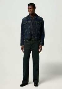 Vyras, apsirengęs tamsiai mėlynu džinsiniu švarku, juoda kapota, juodomis kelnėmis ir juodais batais, stovi prieš vienalytį šviesų foną.