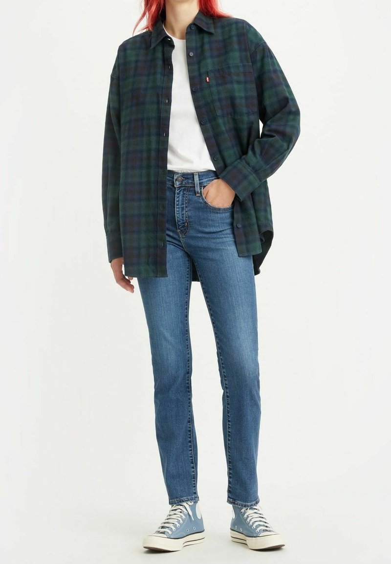 Camisa de cuadros verde y azul marino, camiseta blanca, jeans azules y zapatillas de lona azul claro. La camisa tiene un bolsillo en el pecho y un corte casual.