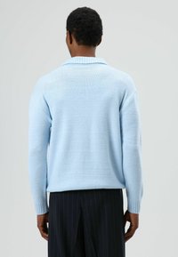 Lichtblauwe gebreide trui met een gestructureerde afwerking, voorzien van een geribbelde kraag en manchetten, gecombineerd met donkere pantalon met fijne strepen.