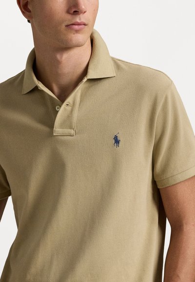 Beige polo särk, mis on valmistatud tekstureeritud kangast, klassikalise kraega, kolme nööbiga ja tumesinise bordüüriga logoga rinna peal.