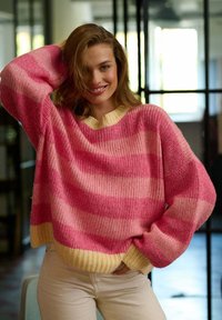 Różowo-kremowy sweter o oversize'owym kroju w paski, z szerokimi rękawami i ribbowanym dołem. Miękka faktura, okrągły dekolt i luźny krój.