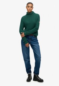 Zielony sweter z golfem i splotem w warkocze, z prążkowanymi krawędziami, w połączeniu z niebieskimi dżinsami o luźnym kroju i czarnymi botkami na sznurówki.