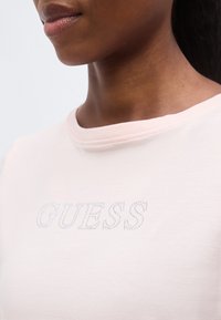 Κοντινό ενός ατόμου που φορά ένα ανοιχτό ροζ πουκάμισο με τη λέξη «GUESS» γραμμένη με μικρές, λαμπερές στρας στο στήθος.