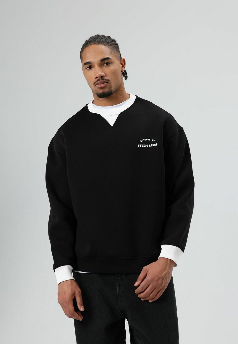 Zwarte sweatshirt met een witte ronde hals en manchetten, voorzien van een V-vormig ontwerp aan de voorkant en witte tekst met de tekst "STUDIO ABYSS." Casual stijl.