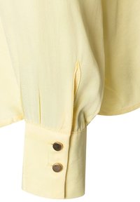 Gele blouse met lange mouwen van zijdezacht stof, manchetten met twee bronzen knopen en geplooide details bij de schouder voor een luchtige uitstraling.