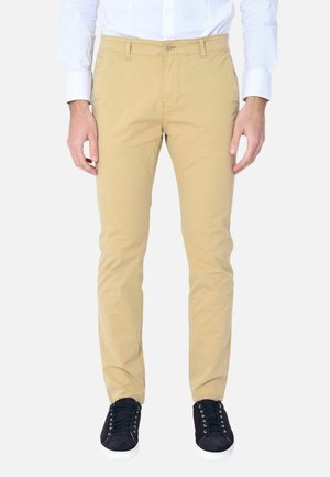 Uomo che indossa pantaloni beige a taglio aderente, camicia bianca a maniche lunghe e scarpe da ginnastica nere, in piedi davanti a uno sfondo bianco.