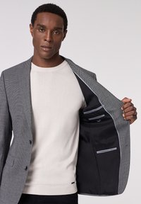 Graue Strukturjacke mit schwarzem Innenfutter, ausgestattet mit Taschen und hellblauen Akzenten, über einem cremefarbenen Pullover getragen.