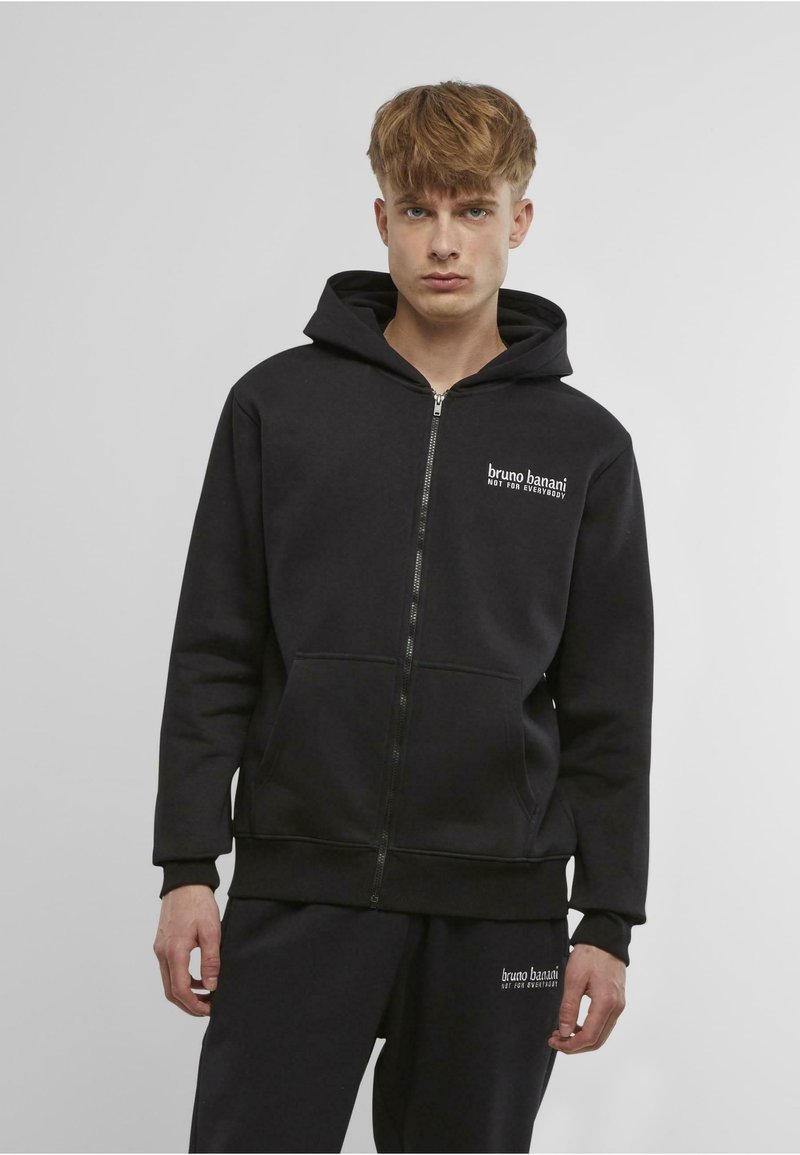 Black zip-up hoodie made of soft fabric, featuring a front pocket and a logo on the chest. Paired with matching black pants.