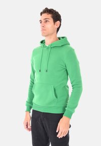 Uomo in piedi che indossa un felpa verde brillante con tasca frontale e pantaloni neri, che guarda leggermente di lato su uno sfondo chiaro e semplice.
