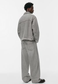 Mlad muškarac okrenut bočno, obučen u labavu sivu traper jaknu i široke traperice s crnim čizmama na bijeloj pozadini.