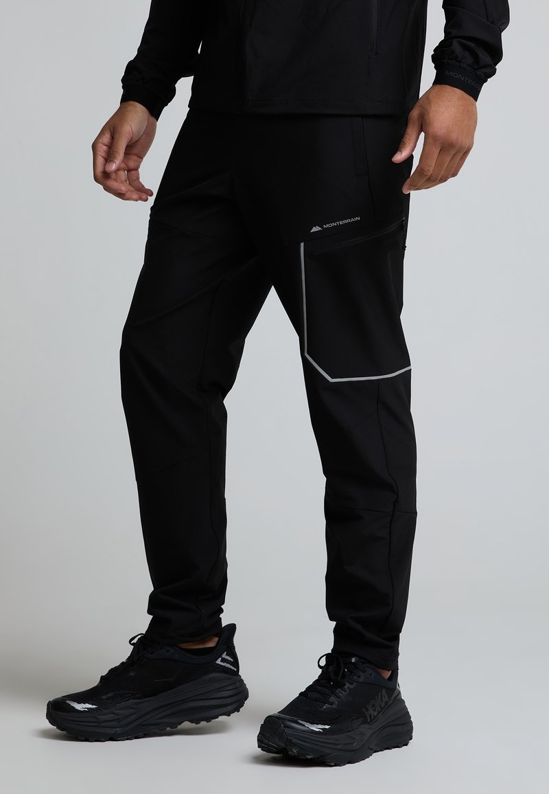 Homme portant un pantalon de plein air noir avec poche zippée et bande réfléchissante, associé à des chaussures de course noires sur fond uni.