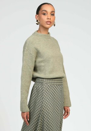 Femme portant un pull en maille vert olive clair, une jupe à carreaux taille haute, de grandes boucles d'oreilles rondes et des bagues aux doigts, debout devant un fond uni.